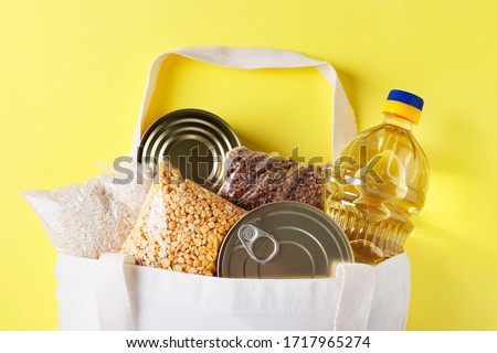 Food delivery, Donation. Textile Bag with food supplies crisis food stock for quarantine isolation period on yellow background. Rice, buckwheat, peas, canned food, vegetable oil, Top view