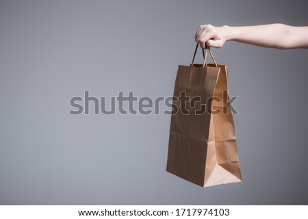 Female hand on a gray background holds out or picks up a paper bag with handles. Advertising shooting for a company with delivery Royalty-Free Stock Photo #1717974103
