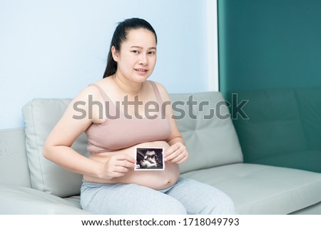 Close up of cute pregnant belly and x ray ultrasound scan of baby. asian young pregnant female relax on couch sofa looking at her child picture in hands sweet. motherhood concept third trimester