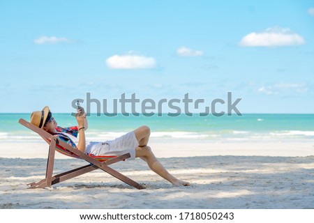 Asian man lying on beach chair using smartphone chatting or online shopping during travel at the beach on summer holiday vacation. People enjoy outdoor lifestyle with gadget device and online network.