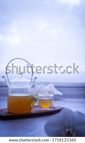 Profile shot of turmeric ginger lemon honey orange tea served in a minimalist glass tea pot cup on wooden platter with beautiful phalaenopsis white orchids in foreground against white glowing window