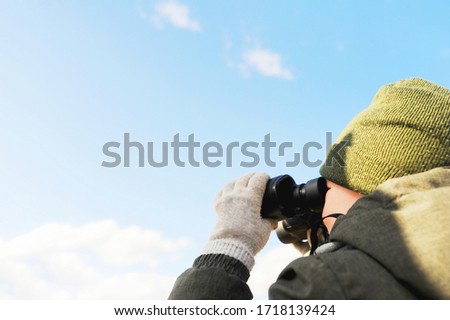 preschool boy in a knitted green hat and a warm green jacket looks at the clear blue sky with clouds in a black binocol