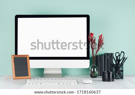 Spring workplace with blank computer monitor, black stationery, books, photo frame, red plant, coffee cup in elegant green mint menthe interior on white wood table.
