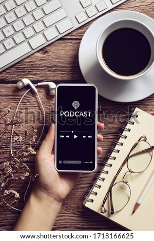 podcast audio content concept. podcast application on mobile smartphone screen on wooden table with coffee cup, earphones, glasses, notebook and pencil. broadcast media Royalty-Free Stock Photo #1718166625