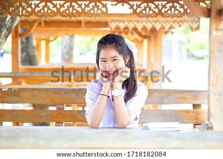 Beautiful Thai female student in a white school uniform sitting on a wooden chair. Asian girl students are smiling on a wooden table.
