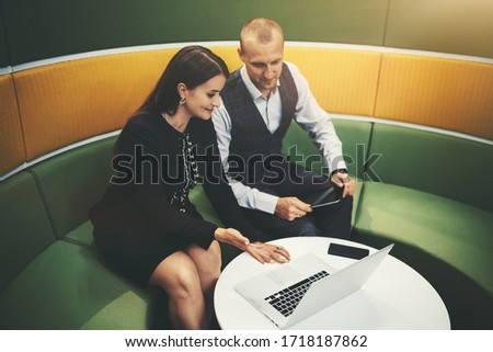 Two business partners having a lively conversation while sitting on a bent sofa of green and yellow colors and using their gadgets: a caucasian businesswoman pointing on the screen of a laptop
