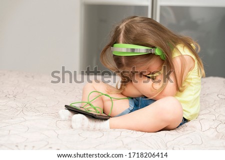 little girl 3-4 years old sitting on the bed in green headphones with a smartphone. Offended pose, face hidden. quarantine and self-isolation in connection with the caronavirus COVID-19