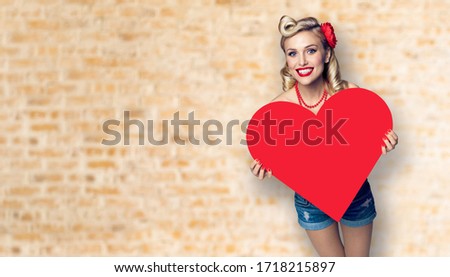 Woman holding red paper heart shape. Pin up girl. Model at retro fashion and vintage concept. Loft style wall background. Copy space for some slogan or sign text. Valentine or Like symbol.