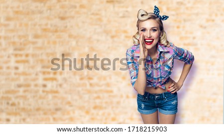 Beautiful happy excited woman holding hand near open mouth. Girl dressed in pin up. Blond model at retro fashion vintage concept, loft style wall background. Copy space area for sign or text.