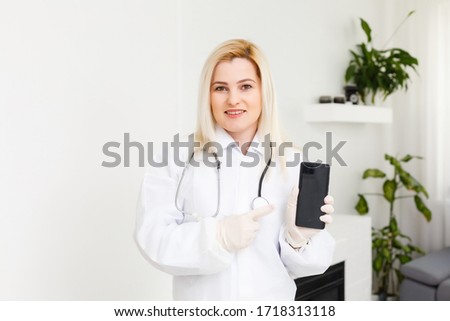 Side profile portrait smiling female doctor, healthcare professional in white lab coat with stethoscope, analyzing data results on mobile smart phone standing in hospital hallway corridor