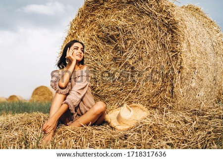A young woman with long hair and in a dress sits near a hay bale. Woman posing smiling and looking at camera.