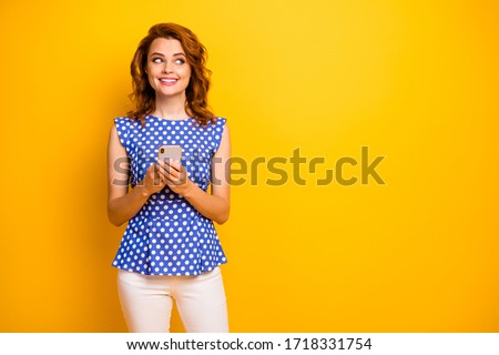 Photo of attractive funny lady blogger hold telephone hands look interested side empty space good mood wear polka-dot blue shirt white pants isolated yellow color background