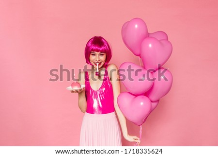 Image of smiling nice woman blowing in party horn while holding balloons and cake isolated over pink background