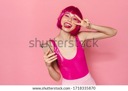 Image of joyful woman in headphone gesturing peace sign and using cellphone isolated over pink background
