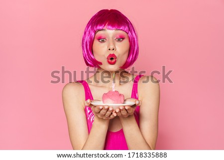 Image of young beautiful woman in wig blowing out candle on cake isolated over pink background
