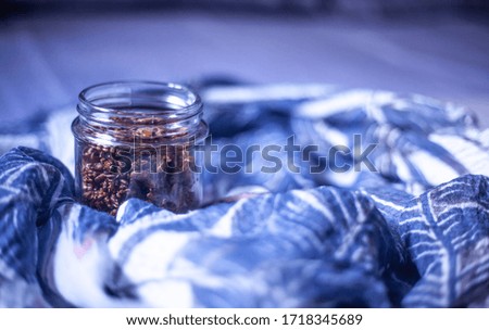 Close up frontal of a glass jar bottle of toasted roasted grilled healthy delicious brown mixed seeds decorated with a beautiful feminine white blue lavender natural leave pattern soft scarf cloth