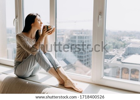 Young woman looking through the window with a city view, sitting on a windowsill, enjoying coffee or tea in the morning. Royalty-Free Stock Photo #1718400526