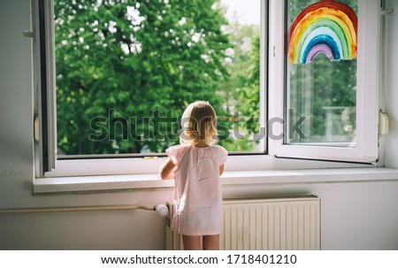 Little girl looks out open window at spring time. Painting rainbow on window. Open window in room with green trees on background. Kids leisure at home. Support during quarantine. Feeling fresh air.