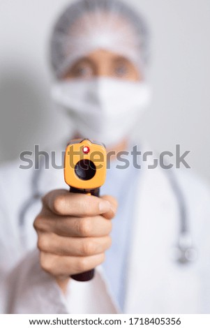 Portrait of a Caucasian female healthcare worker wearing a lab coat, face mask and protective cap against coronavirus, covid 19, holding a non contact thermometer and looking straight into a camera.