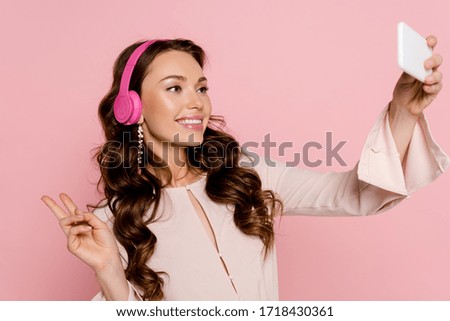 happy girl in wireless headphones taking selfie and showing peace sign isolated on pink