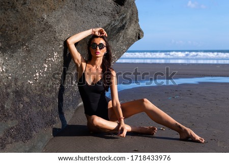 Woman in sunglasses and black swimsuit on black sand beach. Fashionlook woman on beach by ocean. Sunglasses reflect sea and sky. Portrait of woman in sunglasses on Bali.