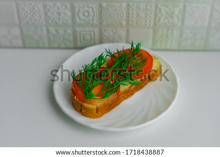 Top view of a piece of bread with butter, slices of red tomato and fresh green dill on white plate on white background