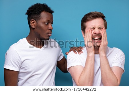African american man calms down his colleague or friend, who has troubles, miserable facial expressions, keeps hands on his shoulders, stands closely against blue background.