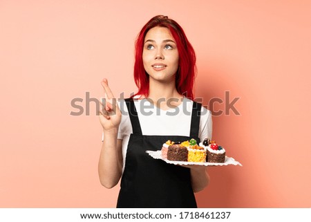 Pastry chef holding a muffins isolated on pink background with fingers crossing and wishing the best