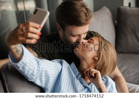 Photo of romantic beautiful nice couple kissing and taking selfie photo on cellphone while sitting on sofa at home
