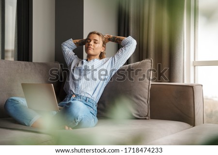 Image of beautiful young relaxed woman resting while sitting with laptop on sofa at living room Royalty-Free Stock Photo #1718474233