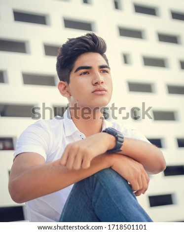 Modern nice young man model in jeans and white shirt sits on floor on a summer outdoor terrace in the city. Stylish attractive hispanic guy modeling outdoors.