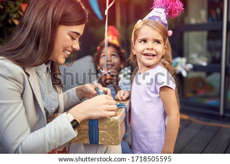young female brunette opening  birthday presents to a little caucasian girl looking away, with afro-american friend of her