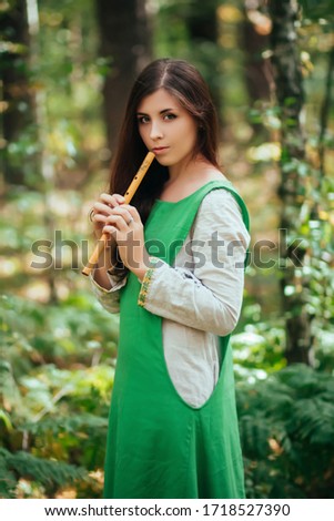 Woman in a green medieval dress plays the wooden flute while standing in a grove against the background of pines. Girl in a gloomy forest plays music on a handmade pipe. Young witch in a gloomy forest