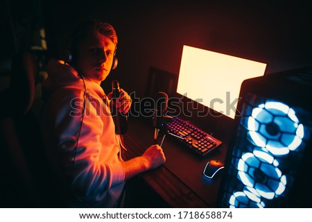 A young man in headphones sits at night at the computer with a beer bottle in his hand and looks into the camera with a serious face. Guy drinks beer and plays video games at night on the computer.