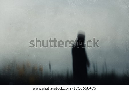 A sinister hooded figure standing in a field, out of focus. With a low camera angle. With Buttercups in the foreground. With a grunge, textured edit. Royalty-Free Stock Photo #1718668495