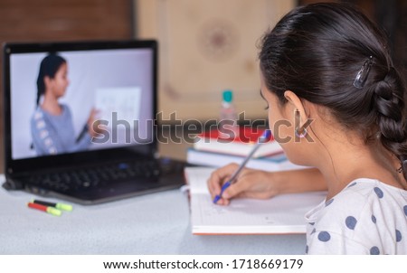 Concept of homeschooling or e-learning, young girl busy in writing by looking into laptop while teacher explaining during covid-19 or coronavirus pandemic crisis Royalty-Free Stock Photo #1718669179