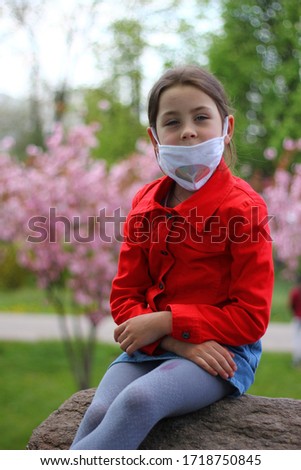 A young girl in a handmade medical mask is sitting on a stone in a red jacket on a background of sakura. Photo taken with daylight.
The picture was taken with selective focus.