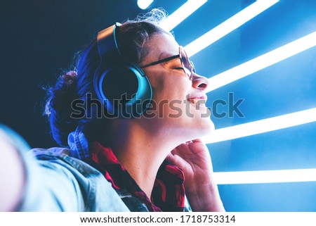 Portrait of modern young woman in headphones making selfie at the blue neon light.