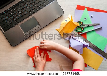 Cute little elementary schoolgirl doing origami fish with folded color paper looking video on laptop, online workshop, kids at-home activity, creativity and distant education Royalty-Free Stock Photo #1718756458