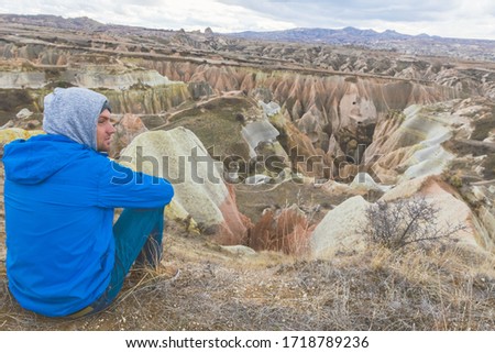 Person with hood and blue jacket sits and calmly wit jot looking around the the landscape of red valley in Cappadocia.