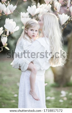 Blonde Mom and daughter on a shiny spring day between magnolia tree flowers dressing white gowns