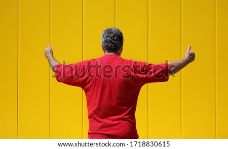 Adult man in red shirt gesturing with thumbs up with yellow metal wall in background