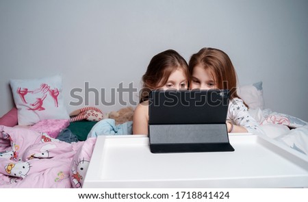 Two little girls sisters lay in the evening in their bed and watched a cartoon on tablets. Cinematic shot. Evening vacation for children. Bedtime tales