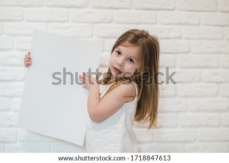 little girl near the white brick wall holding a white empty paper sheet