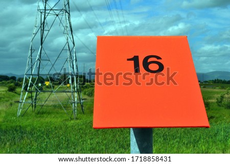Close Up of Red Fire Marker Post 'number 16' in Field beside Steel Power Transmission Pylon 