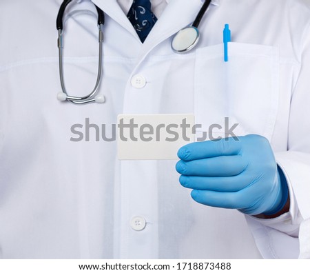 doctor therapist is dressed in a white robe uniform and blue sterile gloves is standing and holding a stack of empty white paper business cards, place for inscription