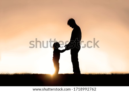 A silhouette of a father and his happy little child are holding hands and smiling at each other as they play outside at sunset.