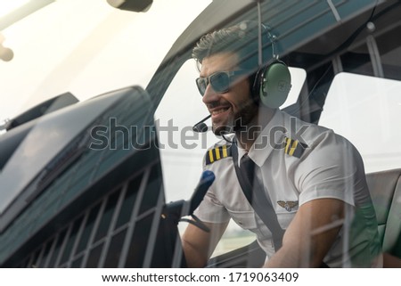 Picture of a male helicopter pilot in his white shirt with tie uniform. He is prepared for his flight. He sits inside a white helicopter with his head phone one ready to take off.
