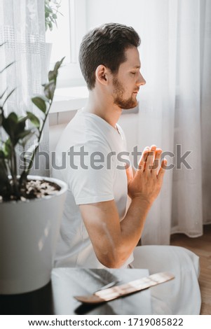 Young man meditating at home on the floor, sitting in the lotus position with closed eyes and folding his hands in Namaste. Expression of tranquility in a health, mindfulness and fitness concept.