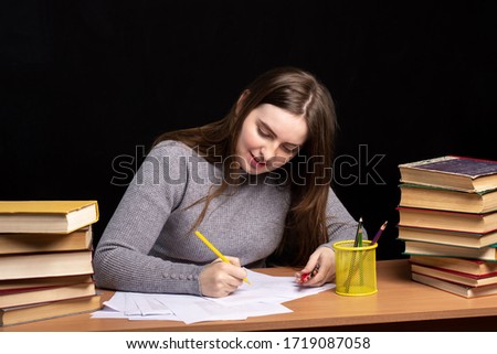 a young girl with long hair writes on paper while sitting at a table with books on a black background. isolate. student preparing for the exam